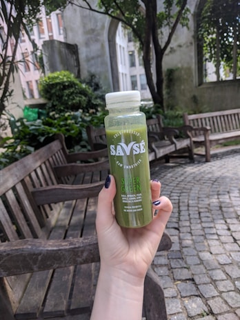 A hand with dark nail polish is holding a bottle of Savse cold-pressed raw smoothie labeled 'Super Green'. The scene is set outdoors in a garden area with wooden benches, stone pathways, and abundant greenery. There is a building in the background with tall windows framed by lush plants and trees.