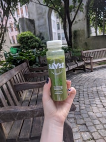 A hand with dark nail polish is holding a bottle of Savse cold-pressed raw smoothie labeled 'Super Green'. The scene is set outdoors in a garden area with wooden benches, stone pathways, and abundant greenery. There is a building in the background with tall windows framed by lush plants and trees.