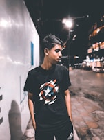 A young person is standing on a dimly lit street at night. They are wearing a black T-shirt with a graphic design on it and Adidas pants. The background features a large, bright wall on the left side and a blurred, bustling cityscape on the right with lights and buildings.