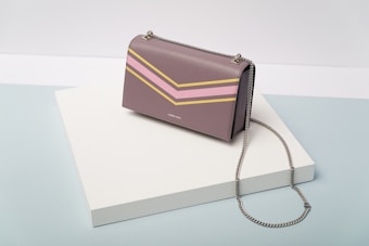 A stylish handbag with a chain strap rests on a white angular platform. The bag has a sleek design featuring chevron patterns in pink and yellow. The backdrop consists of a soft gradient of light blue and cream colors.