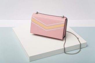 A stylish pink handbag with a metal chain strap is placed atop a white rectangular platform. The handbag features a chevron pattern in white and yellow on its front side. The background consists of light pastel tones in blue and white.
