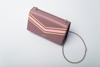 A stylish, mauve-colored handbag with a geometric design featuring pink and yellow lines. It has a metallic chain strap and gold hardware. The bag is placed against a plain, light gray background.