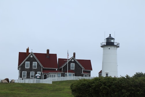 A lighthouse with a black-topped lantern room stands adjacent to a classic wooden house with red roofs. The setting includes a grassy hill in the foreground and an overcast sky. A white picket fence surrounds the house, and the American flag is flying on a pole.