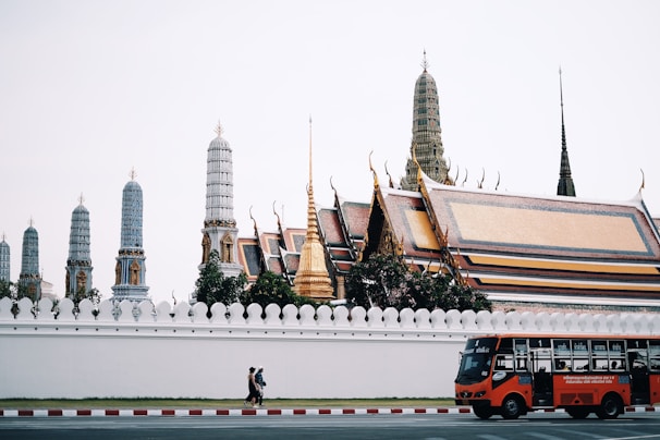A red bus and two people walking past a temple complex with ornate, golden rooftops and tall, intricately designed spires. The temple is enclosed by a white wall, and the architecture features traditional elements.