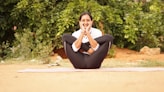 A person is practicing yoga outdoors, seated on a mat with legs crossed, arms wrapped around the knees, and hands resting on the cheeks. The background features green foliage, creating a serene and natural atmosphere.