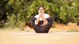 A person is practicing yoga outdoors, seated on a mat with legs crossed, arms wrapped around the knees, and hands resting on the cheeks. The background features green foliage, creating a serene and natural atmosphere.