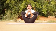 A person is practicing yoga outdoors, seated on a mat with legs crossed, arms wrapped around the knees, and hands resting on the cheeks. The background features green foliage, creating a serene and natural atmosphere.