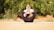 A person is practicing yoga outdoors, seated on a mat with legs crossed, arms wrapped around the knees, and hands resting on the cheeks. The background features green foliage, creating a serene and natural atmosphere.