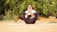A person is practicing yoga outdoors, seated on a mat with legs crossed, arms wrapped around the knees, and hands resting on the cheeks. The background features green foliage, creating a serene and natural atmosphere.