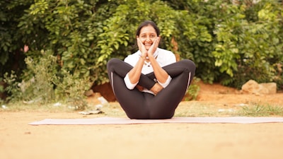 A person is practicing yoga outdoors, seated on a mat with legs crossed, arms wrapped around the knees, and hands resting on the cheeks. The background features green foliage, creating a serene and natural atmosphere.