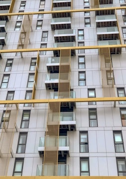 A modern high-rise building facade with a grid-like design. The structure is adorned with numerous small balconies featuring transparent glass railings. Vertical and horizontal yellow beams accentuate the architectural arrangement against the backdrop of white and grey panels. The windows are uniformly spaced and follow a repetitive pattern.