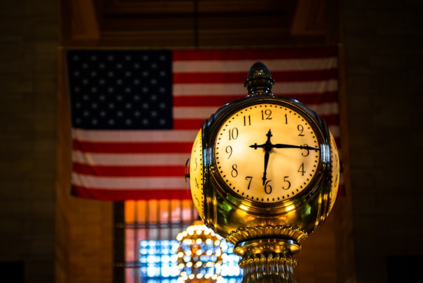 An ornate clock with a golden hue stands prominently in the foreground, displaying the time 10:14. Behind the clock, an American flag hangs inside a dimly lit space, providing a backdrop that adds a sense of patriotism and historical significance.