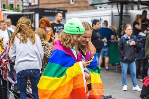 A group of people appear to be gathered at an outdoor event or festival. Two individuals in the foreground are wrapped in a rainbow flag, smiling as they look at a phone. The scene is lively, with several people visible in the background, some holding drinks.
