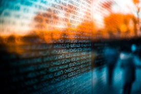 An array of names is inscribed on a reflective surface, possibly a memorial wall. The background features warm, blurred colors reminiscent of a sunset, casting a serene glow. Two blurry figures are faintly visible, adding to the abstract feel.