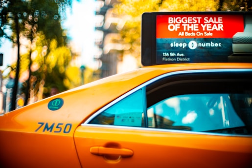 A yellow taxi with a roof-top advertisement for a 'Biggest Sale of the Year' at a retailer is visible. The advertisement highlights a sale on all beds, providing a specific address in the Flatiron District. The taxi's side is marked with a number, and the window reflects some trees and sky. In the background, blurred greenery and city elements create a vibrant urban setting.