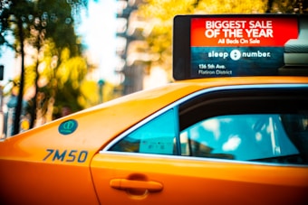 A yellow taxi with a roof-top advertisement for a 'Biggest Sale of the Year' at a retailer is visible. The advertisement highlights a sale on all beds, providing a specific address in the Flatiron District. The taxi's side is marked with a number, and the window reflects some trees and sky. In the background, blurred greenery and city elements create a vibrant urban setting.