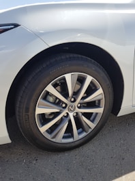 A close-up view of a car wheel featuring a multi-spoke alloy rim with a visible brand logo at the center. The tire tread is detailed and clearly visible, with the brand name 'Michelin' written on the sidewall. The car's body is white, and light is reflecting off its surface.
