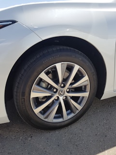 A close-up view of a car wheel featuring a multi-spoke alloy rim with a visible brand logo at the center. The tire tread is detailed and clearly visible, with the brand name 'Michelin' written on the sidewall. The car's body is white, and light is reflecting off its surface.