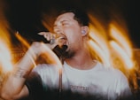 A person passionately singing into a microphone, surrounded by dynamic light trails creating a sense of movement. The individual is wearing a white shirt with an earplug and has a tattoo on the arm. The background is filled with warm, blurred lights.
