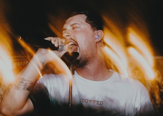 A person passionately singing into a microphone, surrounded by dynamic light trails creating a sense of movement. The individual is wearing a white shirt with an earplug and has a tattoo on the arm. The background is filled with warm, blurred lights.