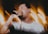 A person passionately singing into a microphone, surrounded by dynamic light trails creating a sense of movement. The individual is wearing a white shirt with an earplug and has a tattoo on the arm. The background is filled with warm, blurred lights.