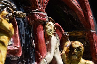 A close-up view of a detailed ceramic sculpture featuring human figures in expressive poses. The foreground figure has an intricate facial expression and is surrounded by textured, multi-colored surfaces. The background includes architectural elements with arches and a variety of carved shapes.