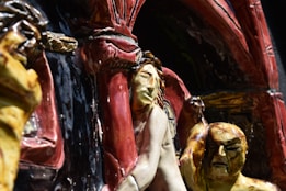 A close-up view of a detailed ceramic sculpture featuring human figures in expressive poses. The foreground figure has an intricate facial expression and is surrounded by textured, multi-colored surfaces. The background includes architectural elements with arches and a variety of carved shapes.