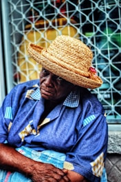 An elderly person wearing a straw hat and a blue patterned shirt sits in front of a metal lattice window. Their expression appears contemplative or thoughtful.