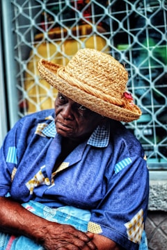 An elderly person wearing a straw hat and a blue patterned shirt sits in front of a metal lattice window. Their expression appears contemplative or thoughtful.