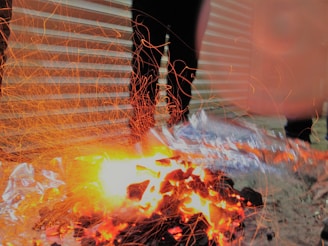 Sparks and embers dance around a small fire, creating dynamic streaks of light against a background of corrugated metal sheets. The fire illuminates the scene with intense yellow and orange hues, contrasting with the shadows of nearby objects.
