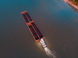 An aerial view of a barge carrying cargo along a wide river. The barge is long, rectangular, and divided into sections, with two sections clearly loaded with dark materials. A small tugboat is pushing the barge forward, creating white waves behind it. The surrounding water appears calm and stretches out towards a distant shoreline.