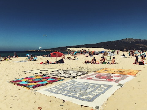 A sandy beach scene featuring people relaxing on colorful towels and blankets. The beach is bustling with activity, with individuals sunbathing and lounging under umbrellas. A clear blue sky stretches above with a few people enjoying kite surfing in the distance. The mountainous landscape provides a scenic backdrop.