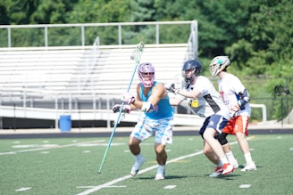 Three lacrosse players are actively engaged in a game on a field. The central player in a blue sleeveless shirt and patterned shorts is moving with a lacrosse stick. Two other players, wearing helmets and team uniforms, are closely defending. The scene is set on a green turf field with bleachers and trees visible in the background.