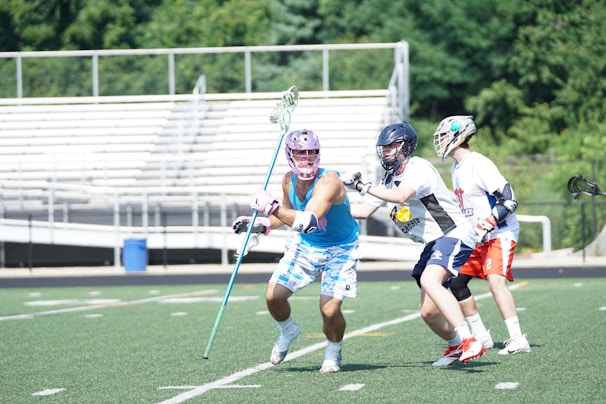 Three lacrosse players are actively engaged in a game on a field. The central player in a blue sleeveless shirt and patterned shorts is moving with a lacrosse stick. Two other players, wearing helmets and team uniforms, are closely defending. The scene is set on a green turf field with bleachers and trees visible in the background.