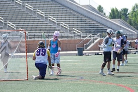 A group of lacrosse players is on a field. One player, wearing a blue and white uniform with the number 99, is kneeling near the goal holding a lacrosse stick. Two other players in white jerseys are standing nearby, wearing protective gear such as helmets and gloves. The field is lined for various sports, and an empty set of bleachers is visible in the background.