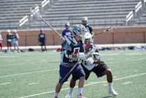Two lacrosse players in action on a field, wearing protective gear and helmets. The player in the foreground holds a lacrosse stick and is closely guarded by another player. The background features empty stadium seating and several other players standing on the sidelines.