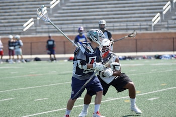 Two lacrosse players in action on a field, wearing protective gear and helmets. The player in the foreground holds a lacrosse stick and is closely guarded by another player. The background features empty stadium seating and several other players standing on the sidelines.