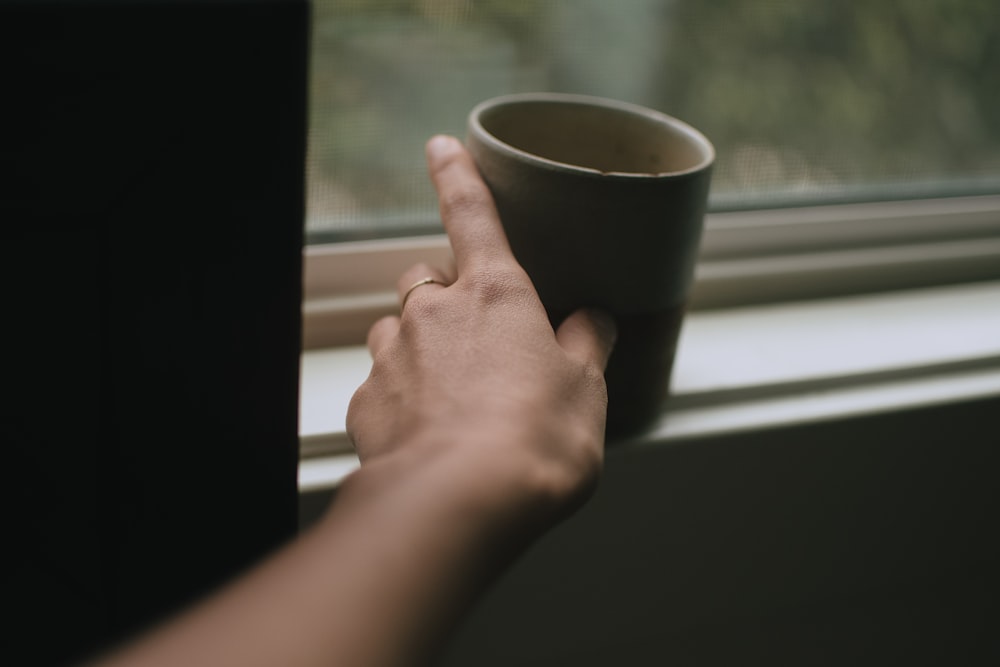 person putting gray ceramic mug near window
