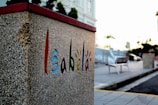 A textured, concrete wall with the word 'Isabela' creatively painted in vibrant colors, including red, blue, green, and orange. The background features a blurred plaza or street scene with a bench, streetlights, and some trees. The light suggests a sunset or sunrise, contributing to a calm atmosphere.