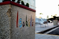 A textured, concrete wall with the word 'Isabela' creatively painted in vibrant colors, including red, blue, green, and orange. The background features a blurred plaza or street scene with a bench, streetlights, and some trees. The light suggests a sunset or sunrise, contributing to a calm atmosphere.