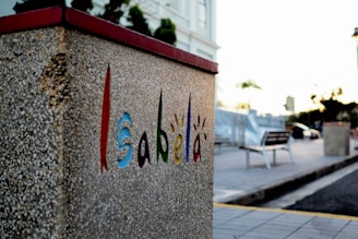 A textured, concrete wall with the word 'Isabela' creatively painted in vibrant colors, including red, blue, green, and orange. The background features a blurred plaza or street scene with a bench, streetlights, and some trees. The light suggests a sunset or sunrise, contributing to a calm atmosphere.