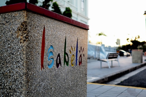 A textured, concrete wall with the word 'Isabela' creatively painted in vibrant colors, including red, blue, green, and orange. The background features a blurred plaza or street scene with a bench, streetlights, and some trees. The light suggests a sunset or sunrise, contributing to a calm atmosphere.