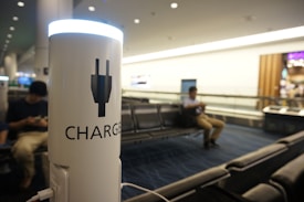 A charging station with a bright top is located in an airport waiting area. The station is labeled with a plug icon and the word 'CHARGE.' The background shows a spacious seating area with a couple of people seated, presumably waiting for their flights. The decor is modern with overhead lighting and a clean, minimalist appearance.