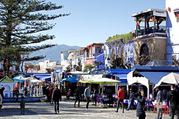 A vibrant marketplace featuring people strolling along a cobblestone street, surrounded by colorful buildings. Numerous stalls covered with awnings display various goods. A large tree provides shade, while the backdrop reveals a mountainous landscape.