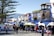 A vibrant marketplace featuring people strolling along a cobblestone street, surrounded by colorful buildings. Numerous stalls covered with awnings display various goods. A large tree provides shade, while the backdrop reveals a mountainous landscape.