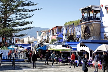 A vibrant marketplace featuring people strolling along a cobblestone street, surrounded by colorful buildings. Numerous stalls covered with awnings display various goods. A large tree provides shade, while the backdrop reveals a mountainous landscape.