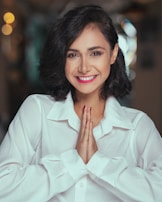 A person with short dark hair is smiling warmly while holding their hands together in a prayer pose. They are wearing a white shirt, and the background is softly blurred with bokeh lights.