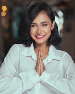 A person with short dark hair is smiling warmly while holding their hands together in a prayer pose. They are wearing a white shirt, and the background is softly blurred with bokeh lights.