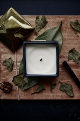 A square, cream-colored candle in a dark container sits on a wooden surface surrounded by dried leaves. A gold, angular object and a small pine cone piece are present nearby. The arrangement is decorated with a dark pen or stick, creating a rustic and natural atmosphere.