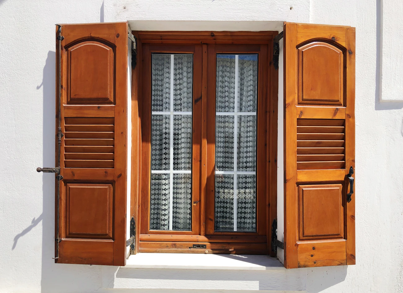 A window with varnished wooden shutters standing open on a whitewashed wall in Megalochori, Santorini, in hard midday sun